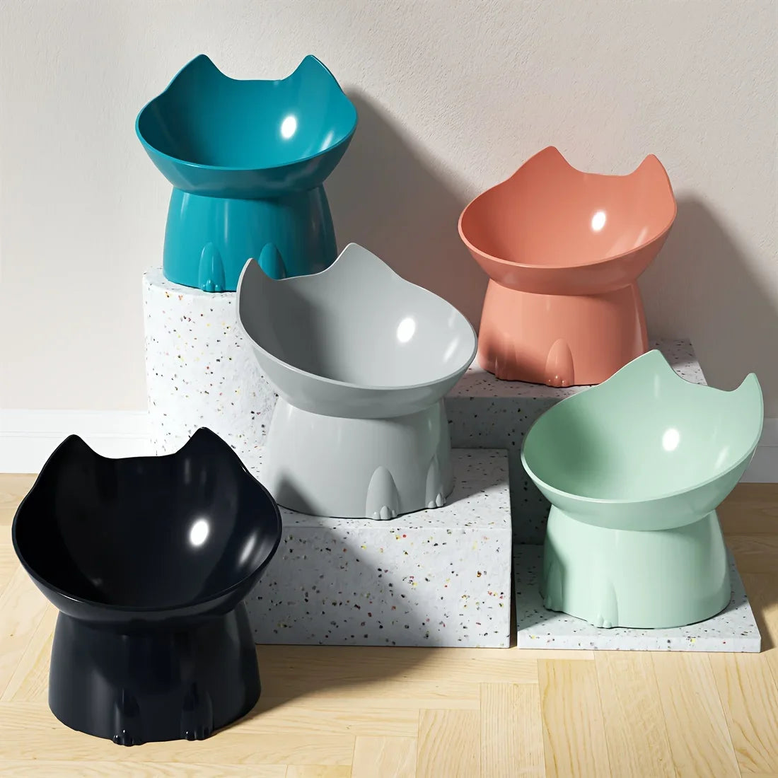 Collection de gamelles surélevées pour chat au design ergonomique en forme de tête de chat, disponibles en noir, gris, bleu turquoise, corail et vert menthe, présentées sur des socles modernes.