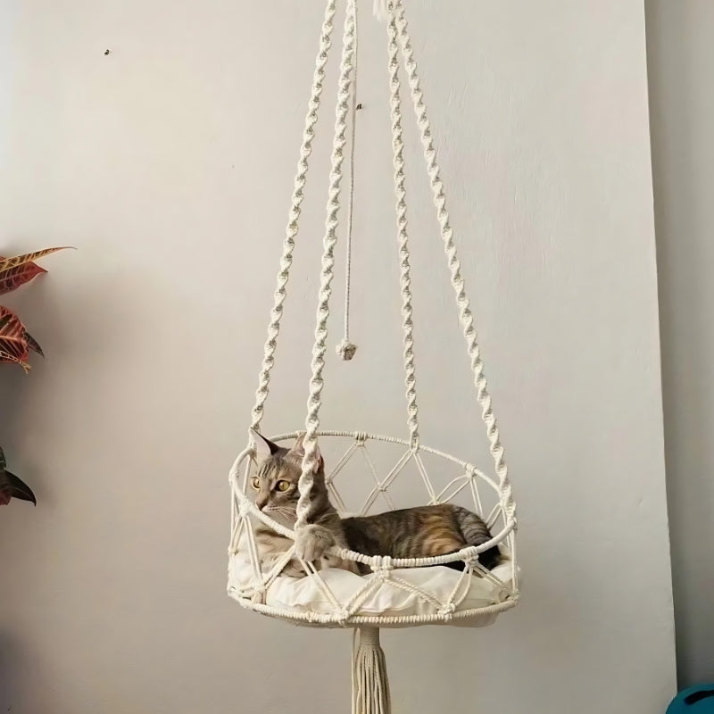 Chat tigré installé dans un hamac suspendu en macramé de coton blanc cassé avec un coussin blanc moelleux, orné de cordes tressées artisanales et d'un pompon décoratif.