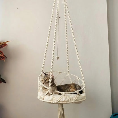 Chat tigré installé dans un hamac suspendu en macramé de coton blanc cassé avec un coussin blanc moelleux, orné de cordes tressées artisanales et d'un pompon décoratif.