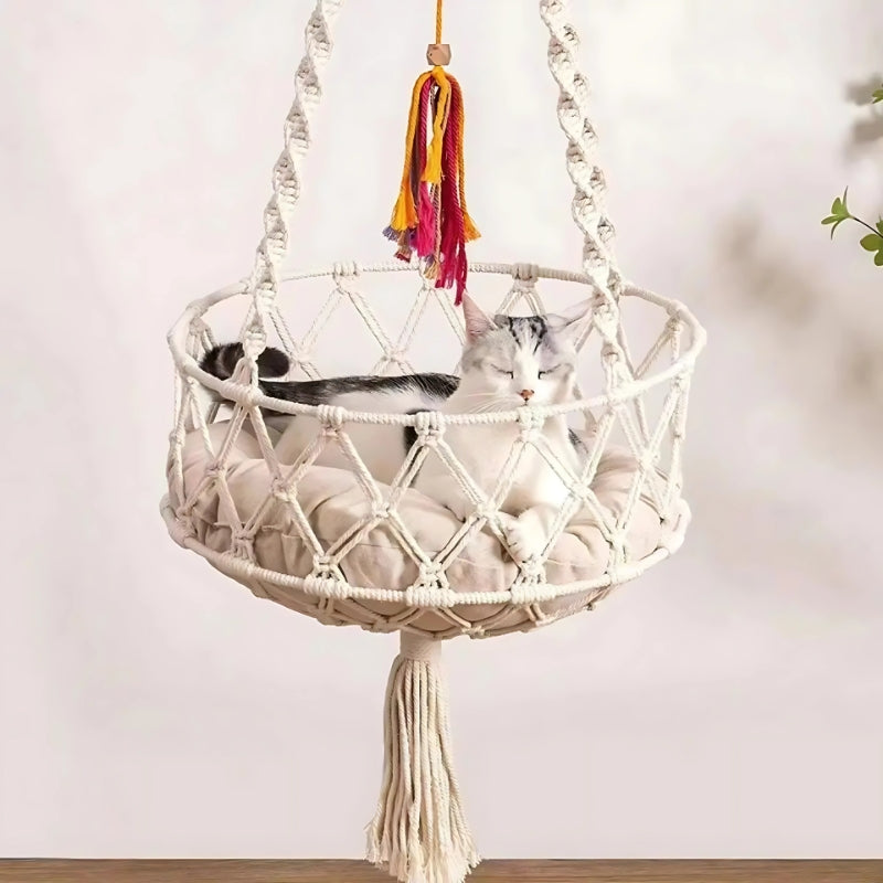 Chat blanc et gris dormant dans un hamac suspendu en macramé de coton blanc cassé avec coussin beige moelleux, orné de cordes tressées et d'un pompon décoratif coloré.
