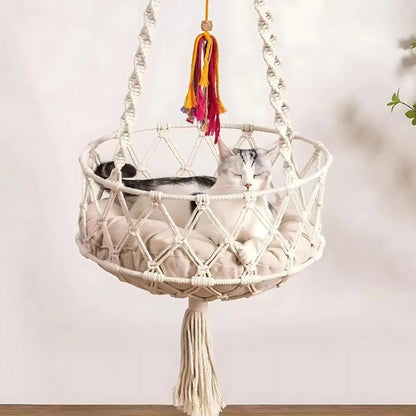 Chat blanc et gris dormant dans un hamac suspendu en macramé de coton blanc cassé avec coussin beige moelleux, orné de cordes tressées et d'un pompon décoratif coloré.