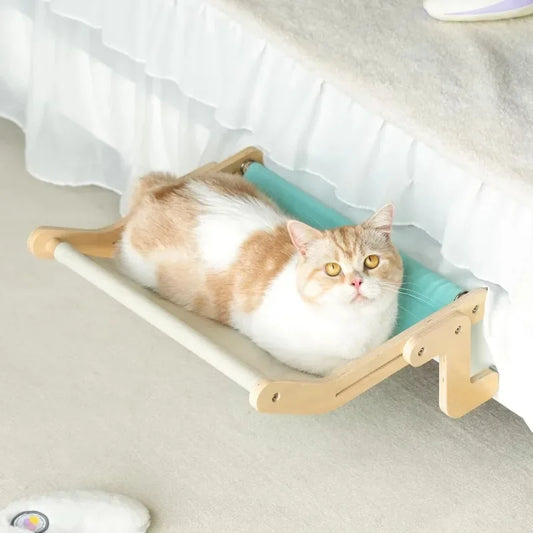 Hamac pour chat en bois clair avec toile turquoise et crème suspendu au bord d'un lit. L'image montre un chat roux et blanc confortablement installé dans son hamac a chat sans ventouse. Lit d'appoint stable, design et robuste pour hamac pour chat de chambre. Qualité premium Les Trésors d'Owka.