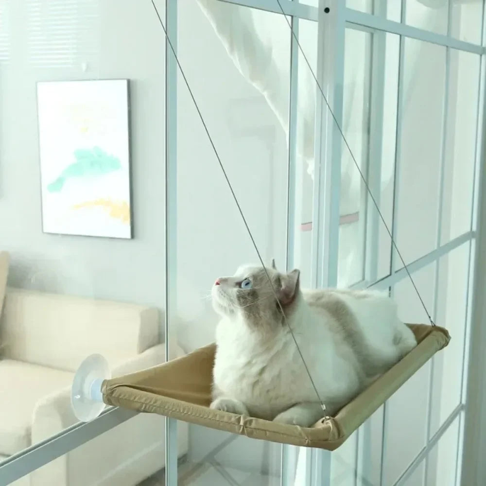Hamac pour chat fixé sur une baie vitrée par des ventouses haute performance. L'image montre un hamac chaton beige au design minimaliste suspendu par des câbles en acier. Lit de fenêtre confortable et discret pour hamac a chat, idéal pour les appartements modernes. Qualité Les Trésors d'Owka.