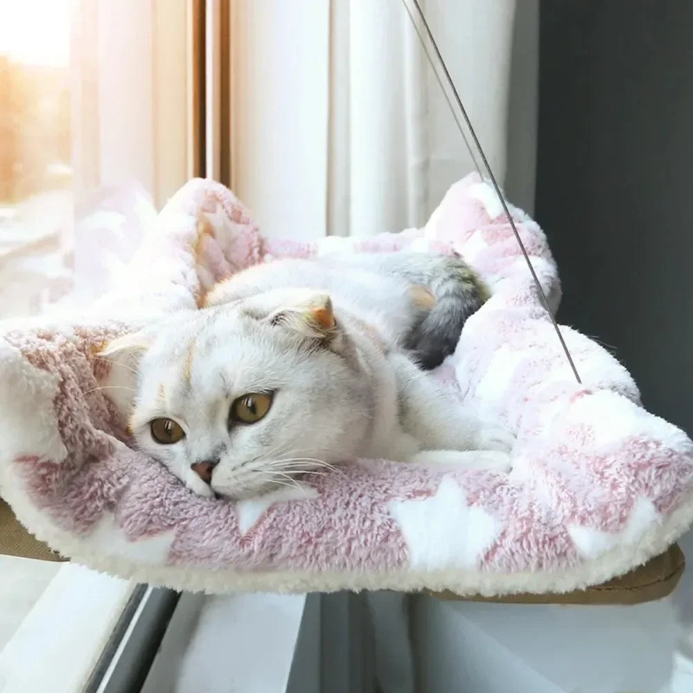 Hamac pour chat de fenêtre avec coussin douillet en peluche rose à motifs étoiles blanches. L'image montre un chaton blanc profitant d'un bain de soleil sur un hamac chaton sécurisé par câbles. Lit suspendu ultra-confortable et design pour hamac a chat. Qualité premium Les Trésors d'Owka.