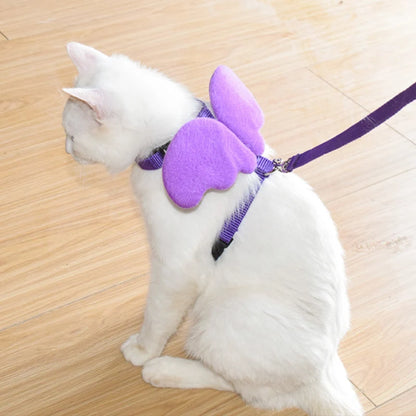 Chat blanc portant un harnais pour chat violet orné d'ailes d'ange en peluche. Ce harnais chat original en nylon avec laisse assortie est ajustable et sécurisé par une boucle clipsable. Un accessoire de type harnais animaux unique alliant mode et sécurité pour des balades féeriques en extérieur.