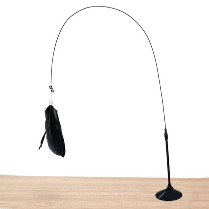 Jouet interactif pour chat avec ventouse de fixation noire posée sur une surface en bois, doté d'une tige flexible noire formant une grande arche et d'un plumeau noir suspendu avec grelot.