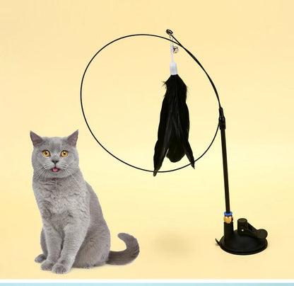 Jouet interactif pour chat composé d'une ventouse noire fixée au sol, d'une tige flexible à ressort et d'un plumeau noir avec grelot, présenté à côté d'un chat gris aux yeux jaunes.