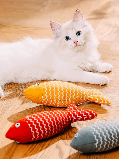 Chaton blanc aux yeux bleus allongé sur un parquet, fixant trois jouets poissons en tissu de couleurs jaune, rouge et gris avec motifs zigzag blancs.