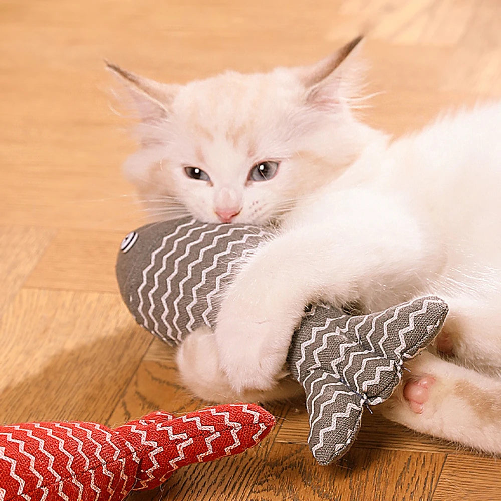 Un chaton blanc et beige allongé sur un parquet joue avec un poisson en tissu gris à motifs zigzag blancs, entouré de deux autres poissons jouets de couleurs jaune et rouge.