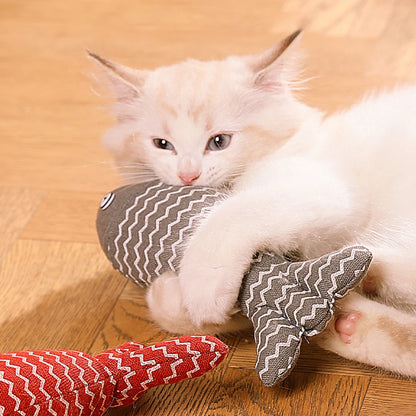 Un chaton blanc et beige allongé sur un parquet joue avec un poisson en tissu gris à motifs zigzag blancs, entouré de deux autres poissons jouets de couleurs jaune et rouge.