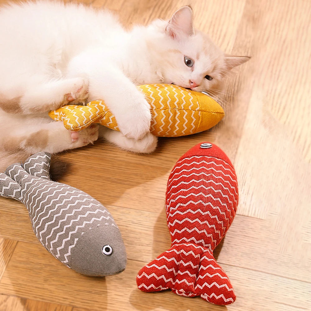 Un chaton blanc et beige allongé sur un parquet joue avec un poisson en tissu jaune à motifs zigzag blancs, entouré de deux autres poissons jouets de couleurs grise et rouge.