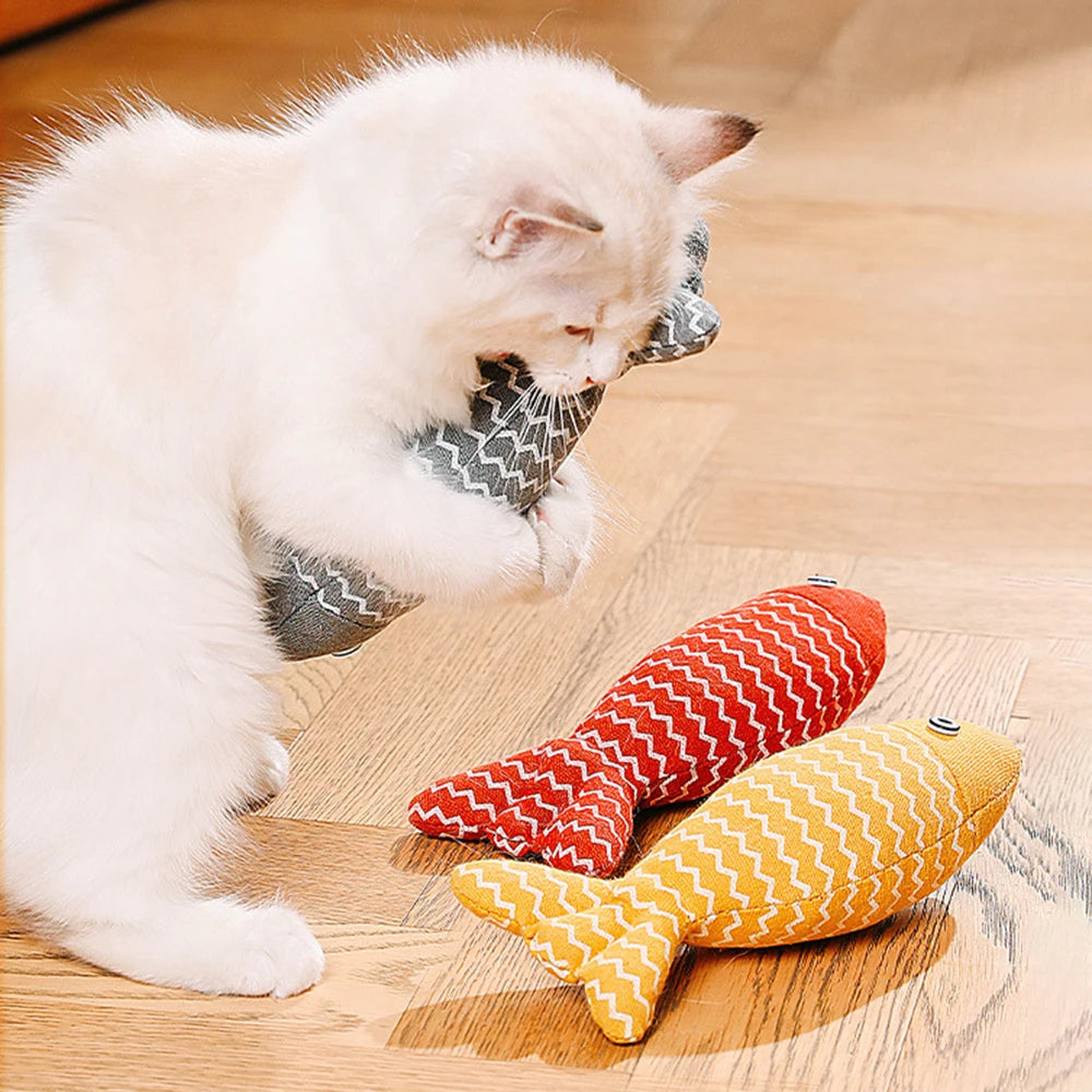 Chaton blanc et beige jouant avec un poisson en tissu gris à motifs zigzag, entouré de deux autres poissons jouets de couleurs jaune et rouge sur un parquet.