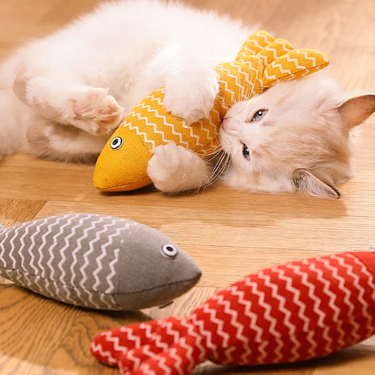 Un chaton blanc et roux allongé sur un parquet joue avec un poisson en tissu jaune à motifs zigzag blancs, entouré de deux autres poissons de couleurs grise et rouge.