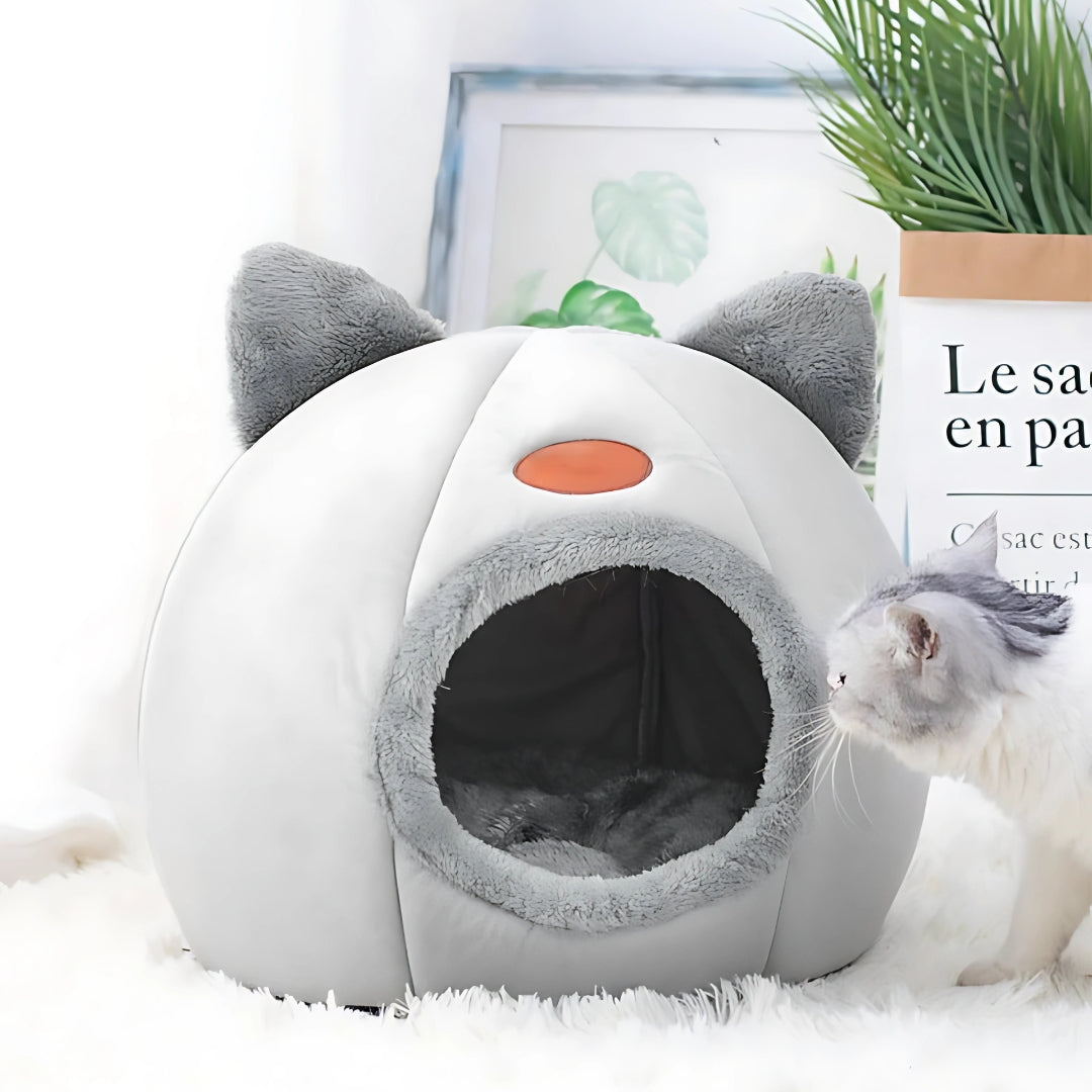 Niche pour chat fermée en forme d'igloo gris et blanc avec oreilles décoratives, intérieur douillet en peluche grise, posée dans un salon moderne à côté d'une plante verte.