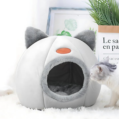 Niche pour chat fermée en forme d'igloo gris et blanc avec oreilles décoratives, intérieur douillet en peluche grise, posée dans un salon moderne à côté d'une plante verte.