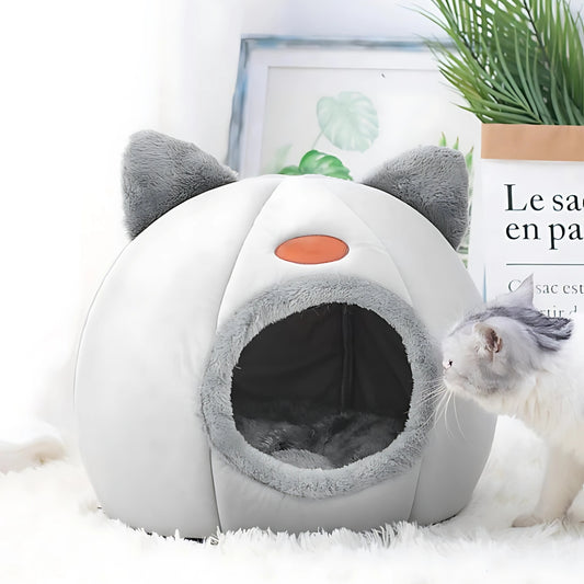 Niche pour chat fermée en forme d'igloo gris et blanc avec oreilles décoratives, intérieur douillet en peluche grise, posée dans un salon moderne à côté d'une plante verte.