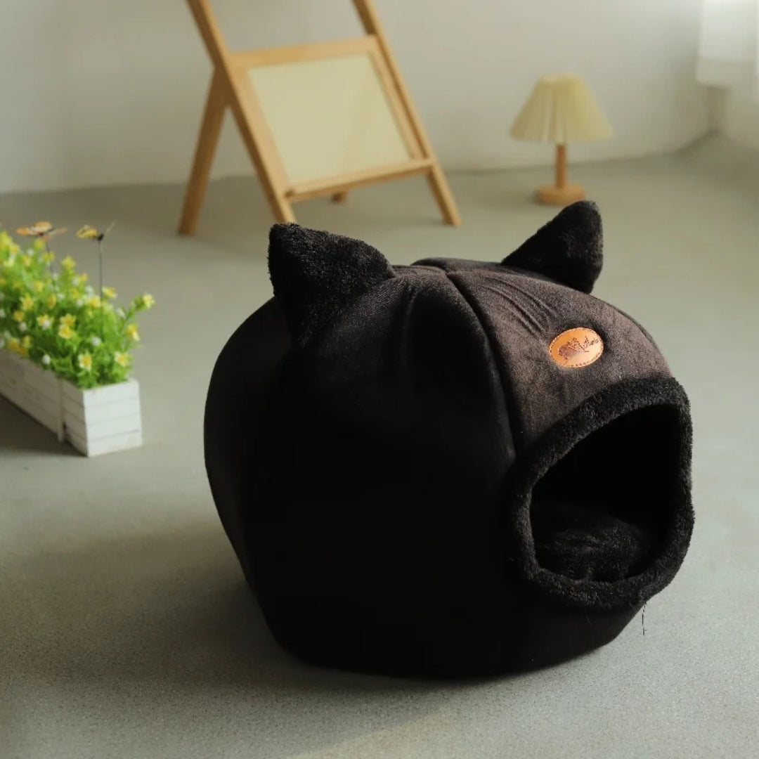 Niche pour chat fermée en forme d'igloo noir profond avec oreilles décoratives, intérieur douillet en peluche noire, posée dans une pièce au design minimaliste avec parquet clair.