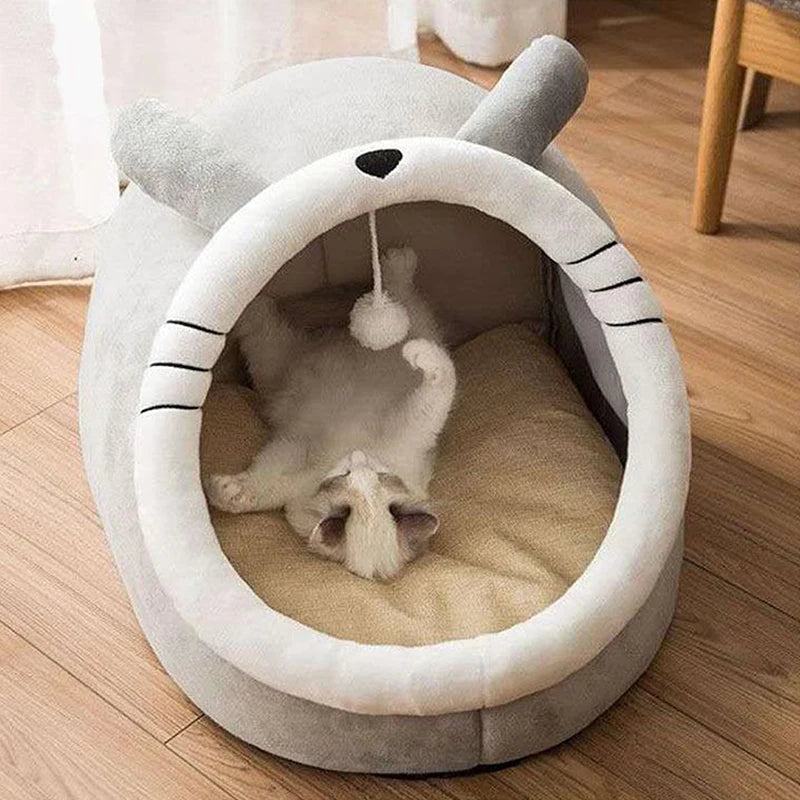 Panier chat en forme de dôme gris avec oreilles et moustaches brodées. L'image montre un chaton couché sur le dos dans ce coussin à chat igloo ultra-moelleux avec pompon de jeu suspendu. Lit fermé protecteur et amusant pour petit animal, parfait comme coussin pour chat cocooning. Qualité Les Trésors d'Owka.