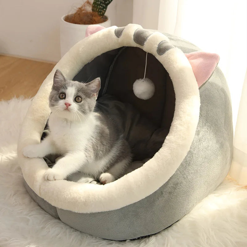 Panier chat en forme de grotte grise avec rebords en peluche blanche et petites oreilles. L'image montre un chaton assis dans ce coussin à chat douillet équipé d'un pompon suspendu. Lit igloo pour petit animal, doux et protecteur, parfait comme coussin pour chat cocooning. Qualité Les Trésors d'Owka.