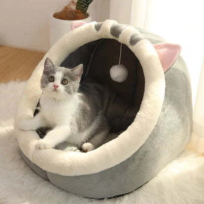 Panier chat en forme de grotte grise avec rebords en peluche blanche et petites oreilles. L'image montre un chaton assis dans ce coussin à chat douillet équipé d'un pompon suspendu. Lit igloo pour petit animal, doux et protecteur, parfait comme coussin pour chat cocooning. Qualité Les Trésors d'Owka.