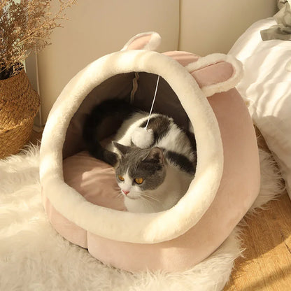 Panier chat en forme de grotte rose poudré avec bordures en peluche blanche et oreilles décoratives. L'image montre un chat installé dans ce coussin à chat igloo ultra-doux équipé d'un jeu pompon suspendu. Lit douillet et protecteur pour petit animal, parfait comme coussin pour chat design. Qualité Les Trésors d'Owka.