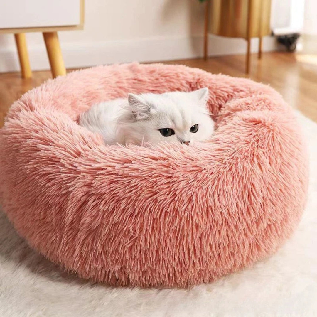Chat blanc niché dans un panier rond apaisant en fausse fourrure rose poudré, montrant les bords surélevés et le rembourrage moelleux pour un confort anti-stress total.