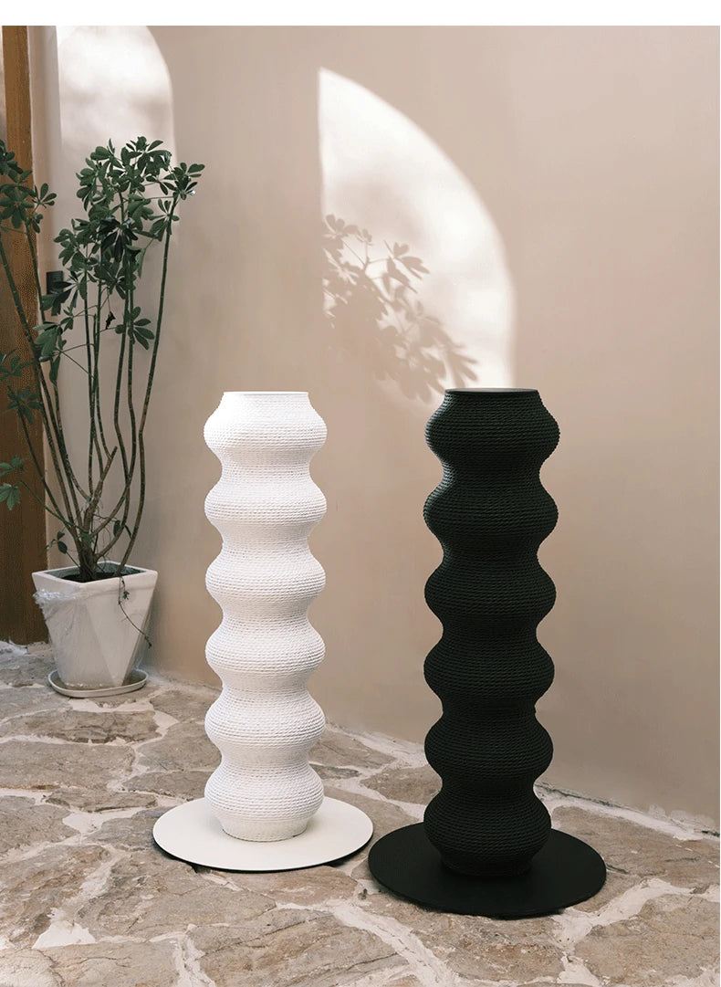 Duo de hauts poteaux à griffer sculpturaux pour chat en forme de colonnes ondulées, présentés en noir et blanc dans un intérieur épuré avec une plante verte.