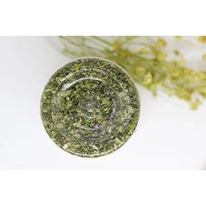 Vue de dessous à travers le fond transparent d'un pot de cataire séchée (Catnip), montrant une texture homogène de feuilles d'herbe à chat finement broyées sans poussière ni résidus.