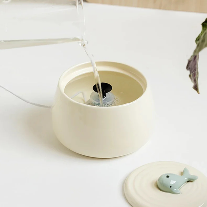 Action de verser de l'eau claire dans le réservoir d'une fontaine pour chat en céramique beige, montrant le filtre interne et le couvercle décoratif avec une petite baleine posé à côté.