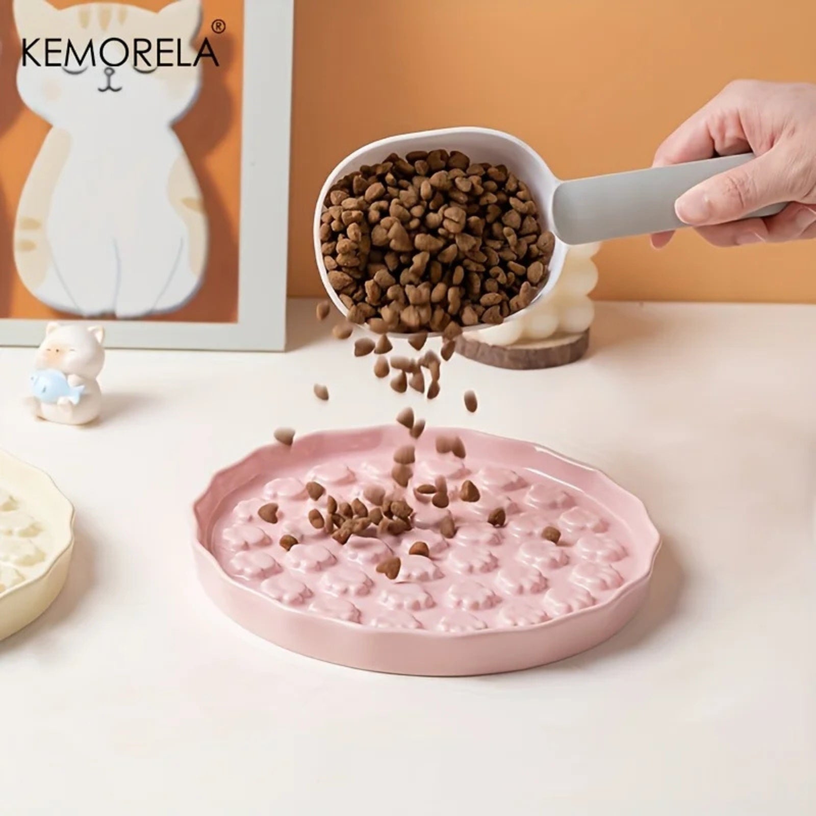 Photo d'une pelle à nourriture versant des croquettes saines dans une gamelle anti glouton chat de couleur rose pastel. Ce plateau ralentisseur en céramique Kemorela, avec son fond texturé de petites pattes de chat en relief, est spécifiquement conçu comme une gamelle glouton pour réguler le débit alimentaire. En arrière-plan, une décoration soignée avec un cadre Kemorela illustrant un chat beige et blanc, renforçant l'univers premium du produit. La chute des croquettes met en avant la profondeur idéale de 