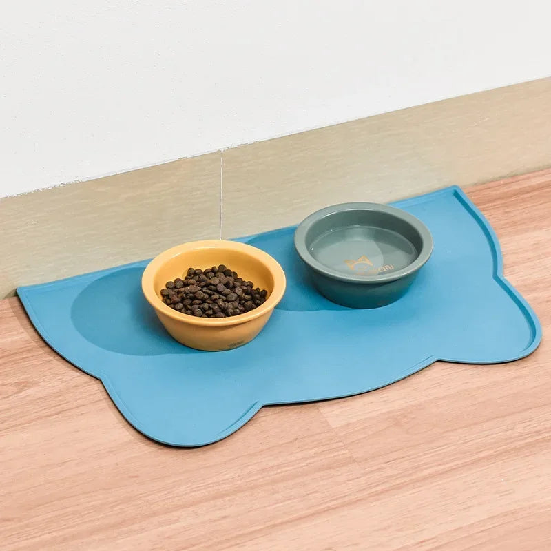 Photographie en situation réelle d'un tapis gamelles chat bleu en silicone posé sur un sol en bois clair. L'image présente le tapis chat design accueillant deux bols : une gamelle jaune pour les croquettes et une gamelle verte pour l'eau. Ce tapis gamelles est doté de bords surélevés intelligents pour contenir les éclaboussures et protéger durablement vos sols de l'humidité. Fabriqué en silicone alimentaire antidérapant, ce tapis chat offre une solution d'hygiène optimale tout en apportant une touche décora