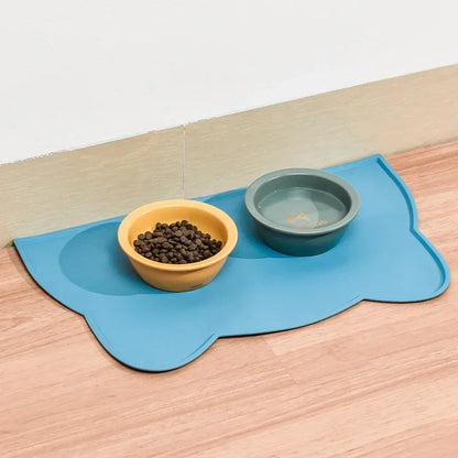 Photographie en situation réelle d'un tapis gamelles chat bleu en silicone posé sur un sol en bois clair. L'image présente le tapis chat design accueillant deux bols : une gamelle jaune pour les croquettes et une gamelle verte pour l'eau. Ce tapis gamelles est doté de bords surélevés intelligents pour contenir les éclaboussures et protéger durablement vos sols de l'humidité. Fabriqué en silicone alimentaire antidérapant, ce tapis chat offre une solution d'hygiène optimale tout en apportant une touche décora