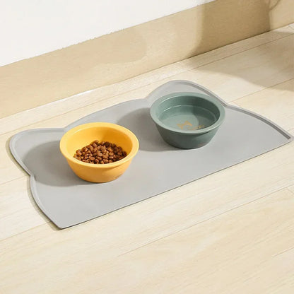 Mise en situation réelle d'un tapis gamelles chat en silicone gris posé sur un parquet clair. L'image présente le tapis chat en forme de tête de félin accueillant une gamelle jaune remplie de croquettes et un bol d'eau vert. Ce tapis gamelles dispose de bords surélevés pour contenir les éclaboussures et protéger durablement vos sols de l'humidité et de la saleté. Sa matière antidérapante et son design épuré en font l'accessoire d'hygiène idéal pour organiser le coin repas de votre animal avec élégance. Une 