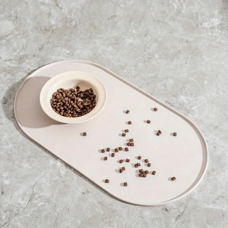 Photographie en situation réelle d'un tapis gamelles chat ovale en silicone beige crème posé sur un sol en marbre gris. L'image montre une gamelle blanche remplie de nourriture avec des croquettes retenues sur la surface du tapis chat, illustrant son efficacité pour protéger vos sols des salissures. Ce tapis gamelles dispose de bords surélevés anti-débordement pour contenir l'eau et les aliments, garantissant une hygiène parfaite du coin repas. Sa matière antidérapante et son design élégant en font l'access