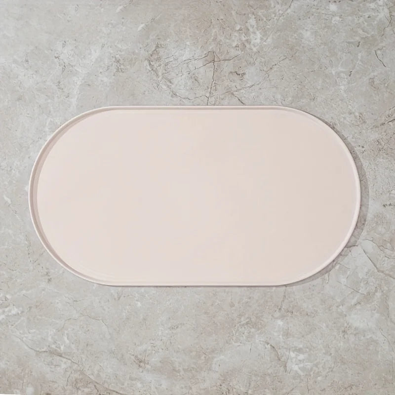 Photographie de face zénithale d'un tapis gamelles chat ovale en silicone blanc crème de la marque Les Trésors d'Owka. Ce tapis chat arbore un design minimaliste avec des bords surélevés anti-débordement pour contenir les projections d'eau et de nourriture, protégeant efficacement vos sols. Fabriqué en silicone alimentaire antidérapant, imperméable et sans BPA, ce tapis gamelles s'intègre parfaitement dans les intérieurs modernes grâce à son coloris ivoire élégant. Sa surface mate et sa facilité de nettoyag