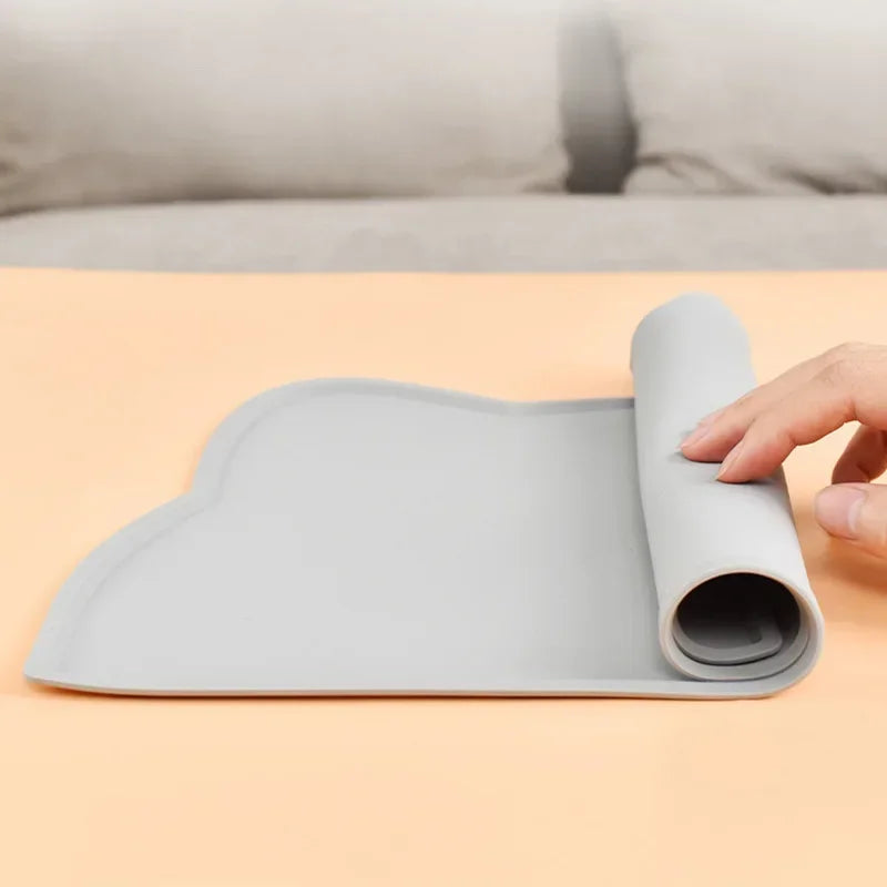Démonstration de la souplesse d'un tapis gamelles chat en silicone gris de la marque Les Trésors d'Owka. L'image montre une main en train d'enrouler le tapis chat pour illustrer sa facilité de rangement et son transport aisé. Ce tapis gamelles de haute qualité est fabriqué en silicone souple et résistant, permettant de gagner de la place tout en conservant ses propriétés antidérapantes et ses bords anti-débordement une fois déplié. Idéal pour les propriétaires nomades ou les petits espaces, ce tapis chat es