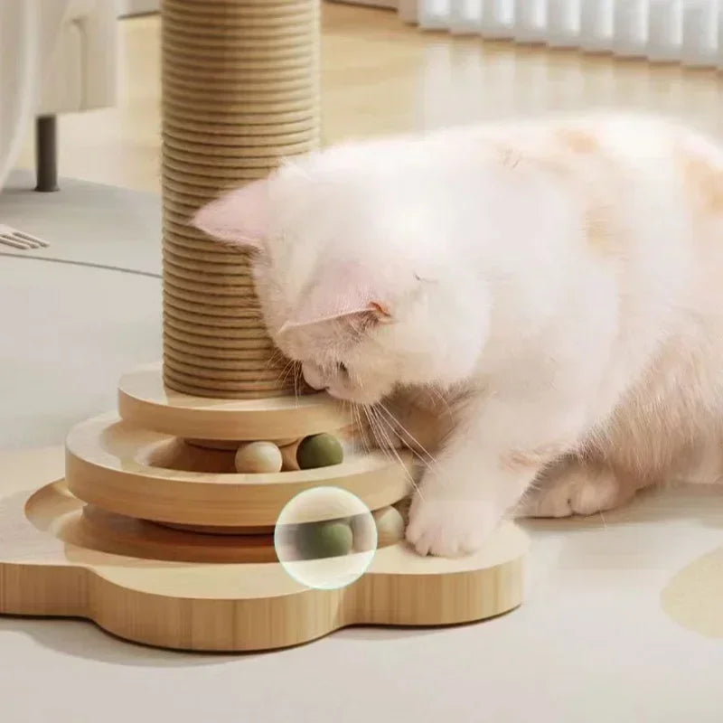 Chat blanc jouant avec un circuit de balles interactif à la base d'une tour de jeu en bois clair, montrant le détail des billes d'herbes à chats et du poteau griffoir en sisal.
